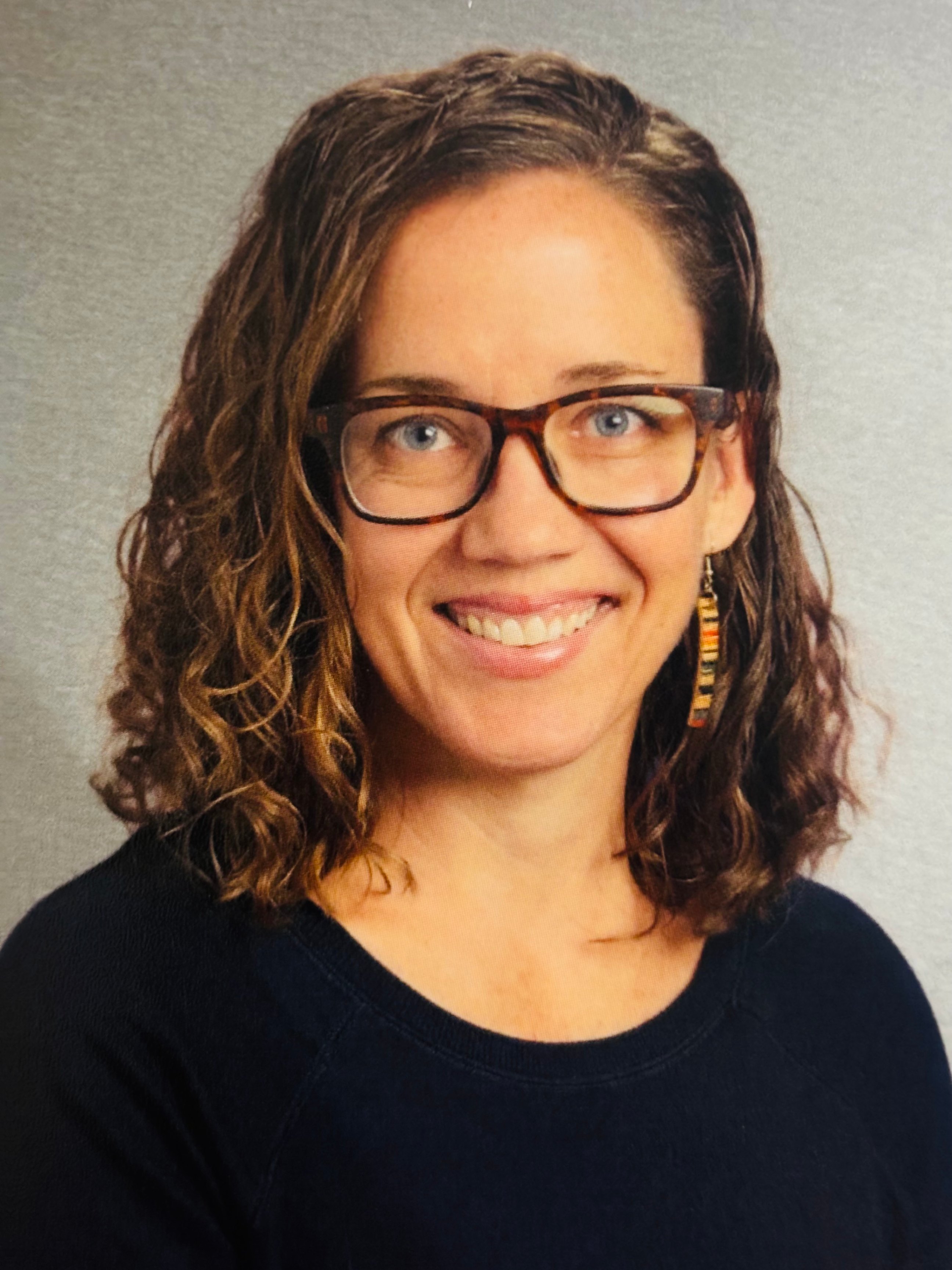 Portrait of a woman outside with wavy hair and glasses and a  big smile. 
